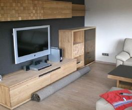 Modernes Wohnzimmer mit Holzmöbeln, Fernseher, Sofa und aufgerolltem Teppich auf dem Boden.