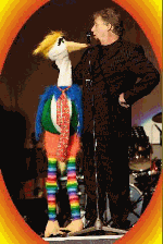 Mann und große, bunte Vogelpuppe miteinander auf einer Bühne; der Mann spricht ins Mikrofon.