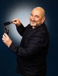 Mann mit Bart, der eine Schere hält und lächelt, trägt ein schwarzes Hemd vor blauem Hintergrund.