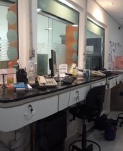 Sala de escritório com computadores, cadeiras, impressoras e janelas de vidro ao fundo.