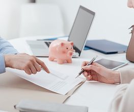 Zwei Personen besprechen ein Dokument am Tisch, mit Laptop und Sparschwein im Hintergrund.