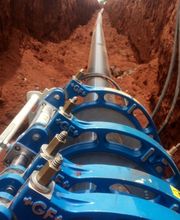 Tubo instalado em vala, segurado por suportes azuis, em terreno de terra avermelhada.