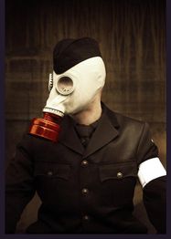 Eine Person in Uniform trägt eine Gasmaske mit rotem Filter, vor dunklem Hintergrund.