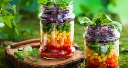 Salada colorida em potes de vidro sobre tábua; inclui milho, pimentão e folhas verdes.