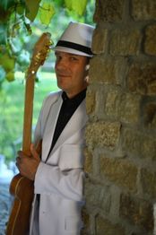 Mann in weißem Anzug mit Hut lehnt an einer Steinmauer, hält Gitarre, umgeben von Blättern.