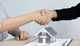 Two people shaking hands over a contract with a small model house in the foreground.
