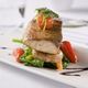Grilled fish with vegetables and garnish on a plate, drizzled with sauce.