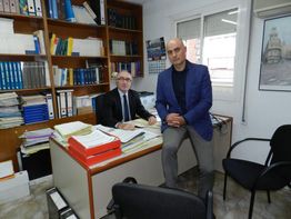 Dos hombres en una oficina, uno sentado en un escritorio y el otro de pie junto a él.