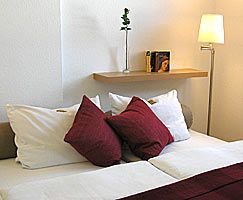 Gemütliches Bett mit weißen und roten Kissen, daneben eine Stehlampe und ein Regal mit Büchern.