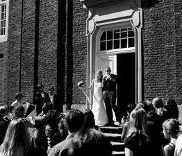 Brautpaar auf Treppe vor einem Gebäude, umgeben von Gästen, schwarz-weiß Fotografie.