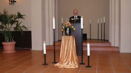 Mann spricht am Rednerpult, umgeben von Kerzen, mit Vase und goldener Decke auf dem Tisch.