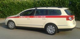 Kremfarbenes Auto mit rotem Streifen und "CityCar" Schriftzug, geparkt vor einer Hecke.
