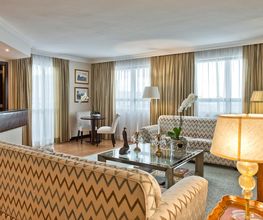 Sala de estar elegante com sofás, mesa de vidro e luminária acesa. Cortinas claras ao fundo.