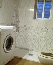 Baño con lavadora, inodoro y ducha. Azulejos blancos y ventana al fondo.