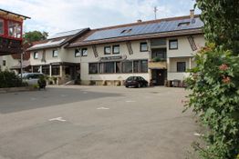 Großes Gebäude mit Solaranlage auf dem Dach, Parkplatz vorne, umgeben von grüner Vegetation.