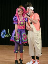 Zwei Clowns in bunten Kostümen auf der Bühne, Clownin hält große blaue Blume.