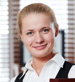 Junge lächelnde Frau in Bluse und Weste vor unscharfem Hintergrund mit Fenster.