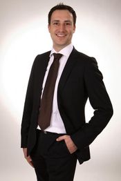 Eine lächelnde Person in Anzug und Krawatte vor einem neutralen Hintergrund.