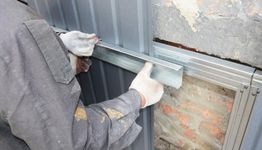 Worker installing metal siding onto a building wall using gloves and tools.