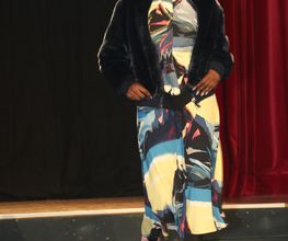 Person in bunter Mode auf Laufsteg, trägt Sonnenbrille und rosa Schuhe, schwarzer Hintergrund.