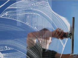 Person reinigt Fensterscheibe mit Abzieher; Schaum bildet Muster, blauer Himmel im Hintergrund.