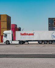Camión transporta contenedor frente a pilas de contenedores en puerto industrial.