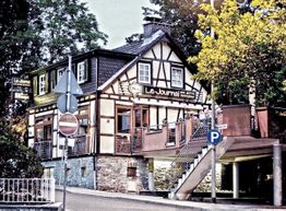 Fachwerk-Restaurant „Le Journal“ mit Treppe und Schildern, umgeben von Bäumen.