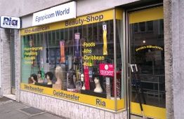 Geschäftsfassade mit Perücken im Schaufenster und Schildern für Geldtransfer und Beauty-Produkte.