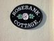 Schwarzes Schild mit Aufschrift "Rosebank Cottage" und einer rosa Blume zwischen grünen Blättern.