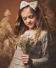 Niña con lazo en el cabello sostiene flores y mira hacia abajo, rodeada de plantas secas.