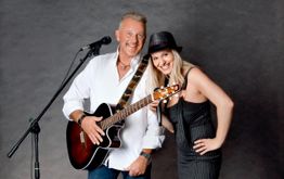 Ein lächelndes Duo mit Gitarre und Mikrofon vor einem grauen Hintergrund.