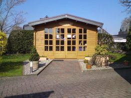 Holzgartenhaus mit bepflanztem Vorgarten, umgeben von Hecken und Bäumen, bei klarem Himmel.