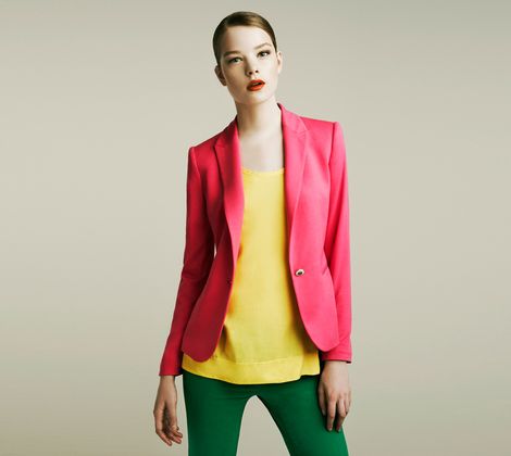 Donna in giacca rosa, top giallo e pantaloni verdi su sfondo grigio chiaro.