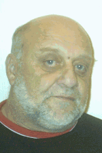 Ein älterer Mann mit Glatze und weißem Bart blickt direkt in die Kamera, hellblauer Hintergrund.