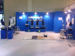 Stand fieristico con pareti blu, macchine esposte e tavoli con sedie.