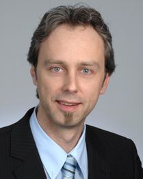 Eine Person im Anzug schaut lächelnd in die Kamera, vor neutralem Hintergrund.
