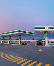 Estación de servicio al atardecer con dos coches repostando y montañas de fondo.