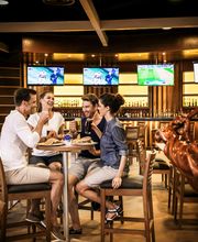 Grupo de amigos comiendo y riendo en un restaurante deportivo con televisores al fondo.