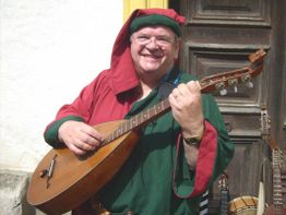 Mann in mittelalterlicher Kleidung spielt eine Mandoline und lächelt vor einer Holztür.