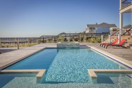 Privater Swimmingpool mit Meerblick, zwei rote Liegestühle, sonniger Tag, modernes Haus im Hintergrund.