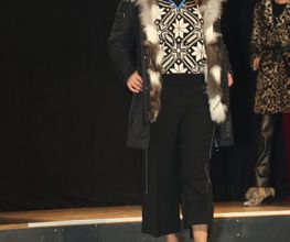 Frau auf Laufsteg mit gemustertem Outfit und Pelzmantel, im Hintergrund Person in Leopardenmuster.