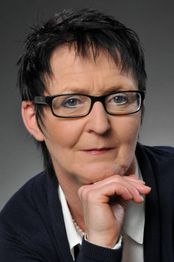 Eine Frau mit Kurzhaarfrisur und Brille blickt in die Kamera, Hand am Kinn.