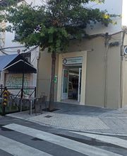 Esquina de calle con tienda, terraza con toldo y árboles bajo cielo despejado.