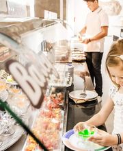 Niña feliz eligiendo postre en un buffet, con un hombre y un chef al fondo.