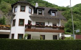 Weißes Haus mit Balkon, umgeben von grüner Hecke und Hügeln im Hintergrund.