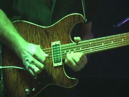 Nahaufnahme von Händen, die eine E-Gitarre mit einer braunen Holzmaserung spielen.