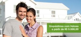 Casal sorrindo em frente a uma casa com anúncio de empréstimos a partir de R$ 5.000,00.