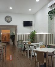 Bar acogedor con mesas, sillas, barra con taburetes y plantas decorativas. Televisor y reloj en pared.