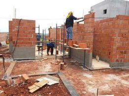 Pedreiros trabalhando em construção de parede de tijolos com andaimes e materiais no chão.