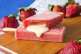 Picolé de morango com recheio cremoso sobre tábua de madeira, rodeado por morangos frescos.